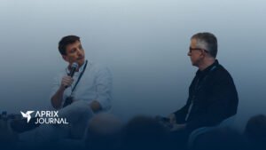 Dois homens em um palco, diante de uma plateia, estão sentados e segurando microfones enquanto conversam, durante um painel do Aprix Connect